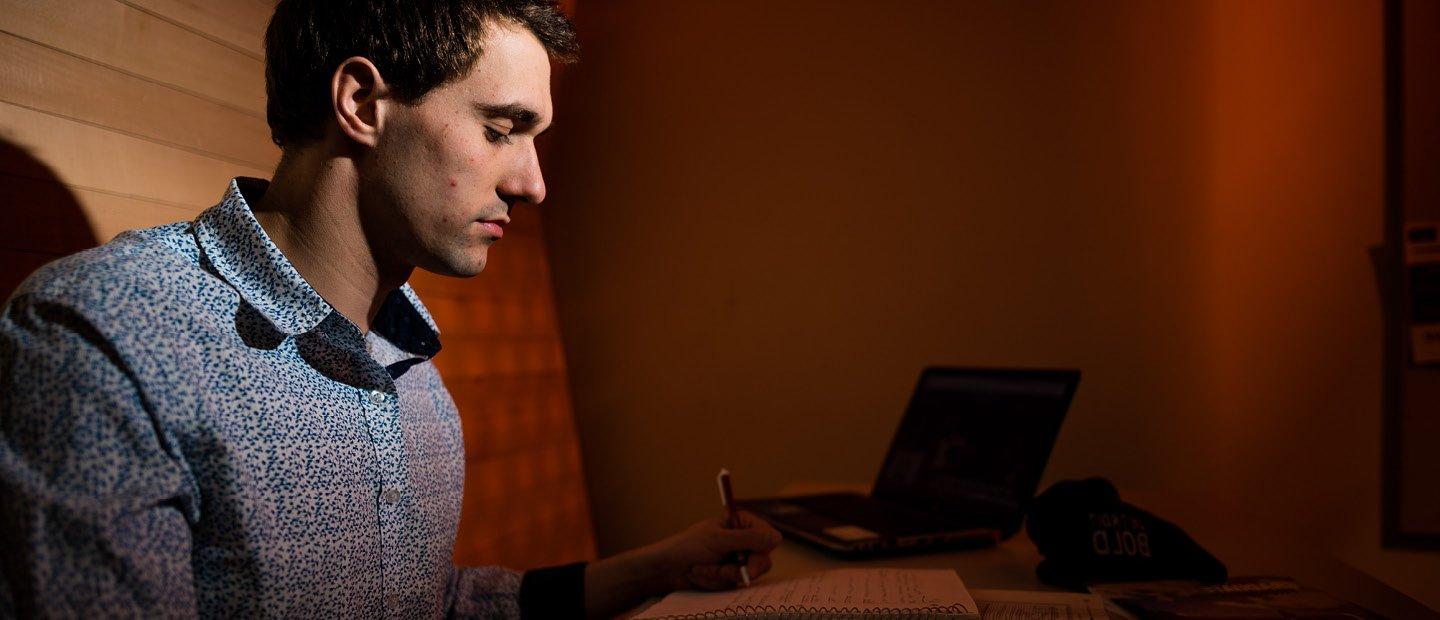 A man sitting in a booth, writing in a notebook with an open laptop next to him.